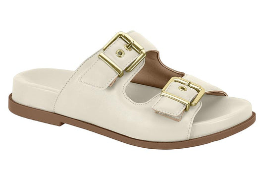 Product shot of a cream-colored leather sandal with two gold buckles and a brown sole showing off its unique design and comfortable features
