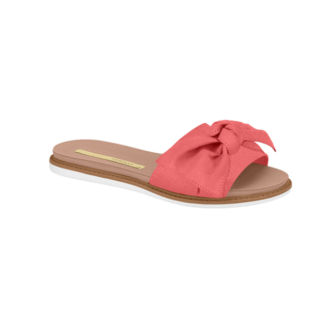 Product shot of a coral-colored Grendha slide sandal featuring a large fabric bow detail a beige insole and a white and brown sole
